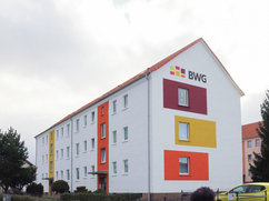 Straßenseitige Ansicht der Wohnhäuser Halberstädter Straße 55 und 57 in Blankenburg mit frisch sanierter weißer Fassade. Farbige Akzente in Rot, Gelb und Orange schmücken die Giebelseite, darüber das BWG-Logo. Der Eingangsbereich ist mit einem orangefarbenen Fassadenstreifen betont.