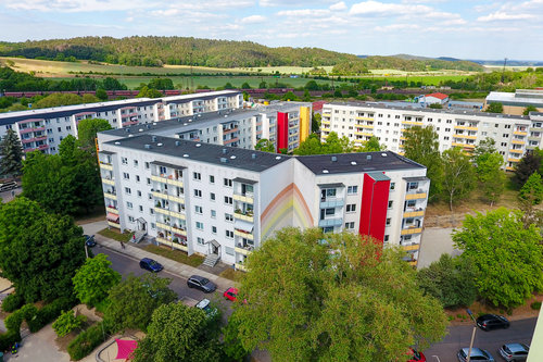 Luftaufnahme des Wohnquartiers Regenstein in Blankenburg mit mehreren sanierten Mehrfamilienhäusern in heller Fassadengestaltung, teilweise mit roten Akzenten und Regenbogenmotiv. Im Hintergrund grüne Hügel, Felder und ein Zug auf der Bahnstrecke.
