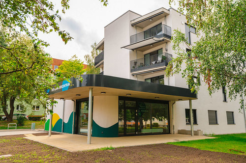 Ansicht des Eingangsbereichs der neuen Sozialstation im Quartier Regenstein. Zu sehen ist ein moderner, überdachter Zugang mit farbenfroher Fassadengestaltung in Grün-, Gelb- und Weißtönen. Im Hintergrund das mehrstöckige Wohngebäude mit Balkonen, umgeben von Bäumen und Grünflächen.