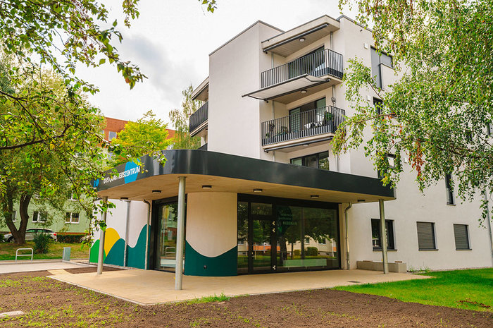 Ansicht des Eingangsbereichs der neuen Sozialstation im Quartier Regenstein. Zu sehen ist ein moderner, überdachter Zugang mit farbenfroher Fassadengestaltung in Grün-, Gelb- und Weißtönen. Im Hintergrund das mehrstöckige Wohngebäude mit Balkonen, umgeben von Bäumen und Grünflächen.