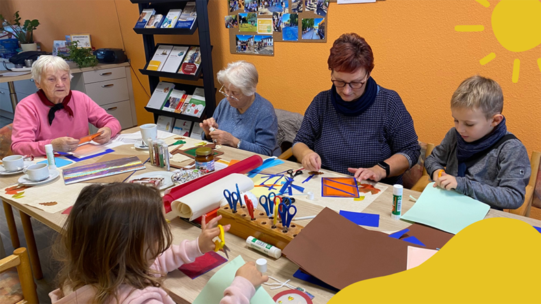 Senioren und Kinder sitzen zusammen an einem Basteltisch mit buntem Papier, Stiften und Scheren – ein generationenübergreifendes Kreativangebot in freundlicher Atmosphäre.