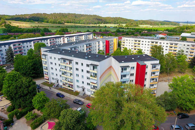 Luftaufnahme des Wohnquartiers Regenstein in Blankenburg mit mehreren sanierten Mehrfamilienhäusern in heller Fassadengestaltung, teilweise mit roten Akzenten und Regenbogenmotiv. Im Hintergrund grüne Hügel, Felder und ein Zug auf der Bahnstrecke.