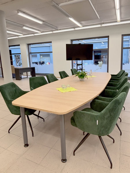 Acht grüne Stühle stehen an einem langen Tisch. Außerdem steht in dem Raum ein Fernseher