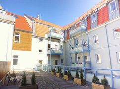 Innenhof des Wohnkomplexes Lange Straße 42 / Tränkestraße 21 in Blankenburg. Gepflegter Bereich mit Sitzbank, Pflanzkübeln und Holzsteg. Umgeben von sanierten Gebäudeteilen mit roten Dachgauben, Balkonen und modernen Metallgeländern.