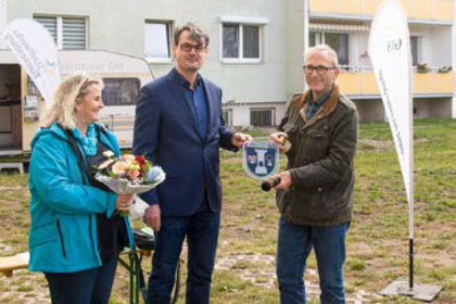 Übergabe eines Wappenschilds auf einer Freifläche: Drei Personen bei feierlichem Anlass vor einem Wohnblock – mit Blumenstrauß, Mikrofon und Info-Fahne der Wohnungsbaugesellschaft