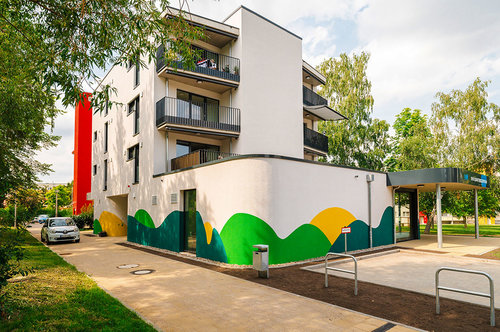 Fotografie des fertiggestellten Neubaus im Quartier Regenstein: Ein modernes, mehrstöckiges Wohngebäude mit Balkonen, bunter Fassadengestaltung in Grün-Gelb-Tönen und einem überdachten Eingangsbereich. Im Vordergrund befinden sich barrierefreie Zugänge, Fahrradbügel und ein kleiner Vorplatz.