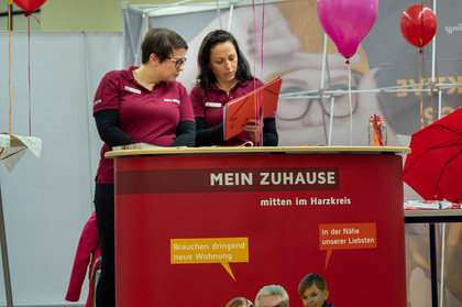 Zwei Mitarbeiterinnen der BWG am Messestand im Harzkreis im Gespräch mit Infomaterial, Werbebanner mit der Aufschrift „MEIN ZUHAUSE mitten im Harzkreis“ und roten Dekoelementen im Hintergrund
