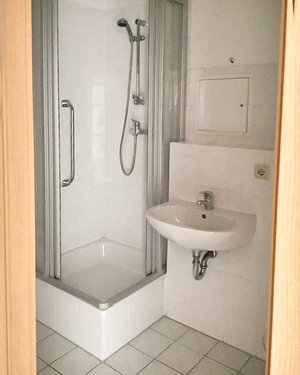 Das Bild zeigt ein Badezimmer mit weißen Fliesen und einer Dusche mit Ecktür. Daneben befindet sich ein Waschbecken.