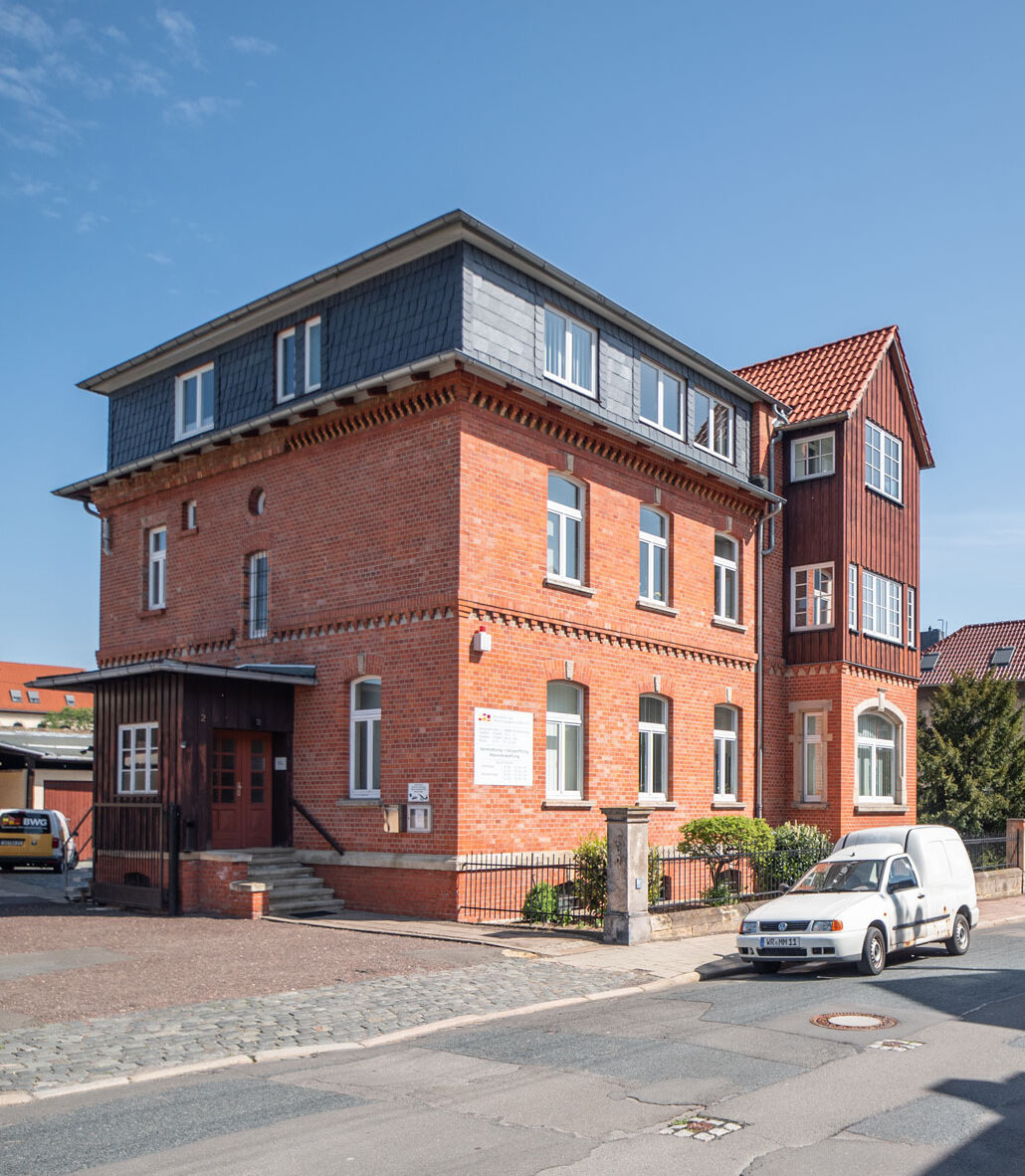Backstein-Gebäude mit ausgebautem Dachgeschoss und Erker, davor ein geparktes weißes Auto auf einer Straße.