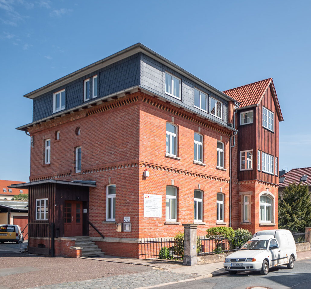 Backstein-Gebäude mit ausgebautem Dachgeschoss und Erker, davor ein geparktes weißes Auto auf einer Straße.