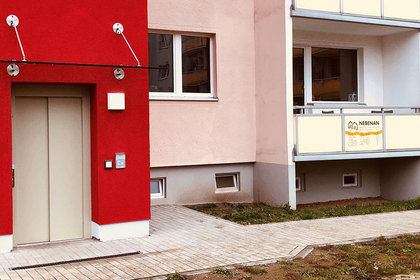 Neubau mit roter und rosafarbener Fassade, Aufzugseingang und Balkon mit dem Schild „NEBENAN“ – zeigt modernen, barrierefreien Wohnraum im Quartier.