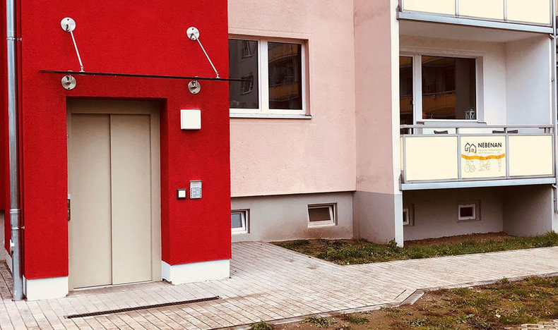 Neubau mit roter und rosafarbener Fassade, Aufzugseingang und Balkon mit dem Schild „NEBENAN“ – zeigt modernen, barrierefreien Wohnraum im Quartier.