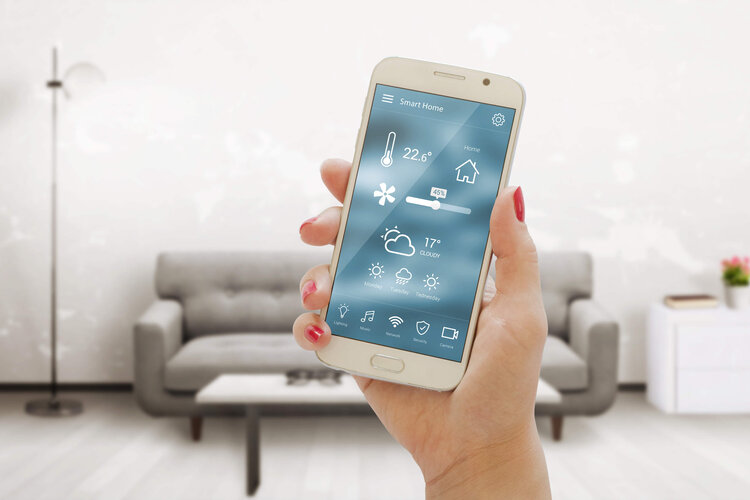 Smartphone mit Smart-Home-App zur Steuerung von Temperatur, Licht und Sicherheit – moderne Wohntechnologie im Wohnzimmer mit grauem Sofa im Hintergrund