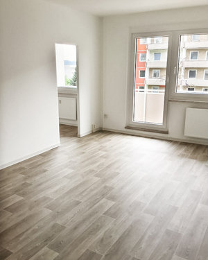 Das Foto zeigt ein Zimmer mit Balkon und weißen Wänden. Auf dem Boden ist grau-braunes Parkett zu sehen.