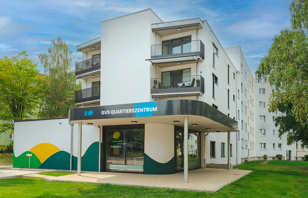 Frontalansicht des modernen GVS Quartierszentrums in Blankenburg mit weißer Fassade, Balkonen und überdachtem Eingangsbereich. Am Gebäude befindet sich ein Schild mit der Aufschrift „GVS Quartierszentrum“. 