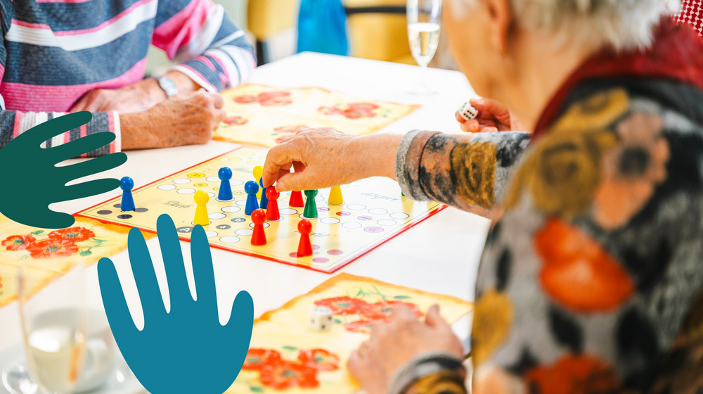 Seniorinnen und Senioren spielen gemeinsam an einem Tisch das Brettspiel „Mensch ärgere dich nicht“. Die Szene zeigt bunte Spielfiguren, fröhliche Tischdekoration mit Blumenmotiven und grafische Hand-Symbole im Vordergrund, die für Miteinander und Gemeinschaft stehen.
